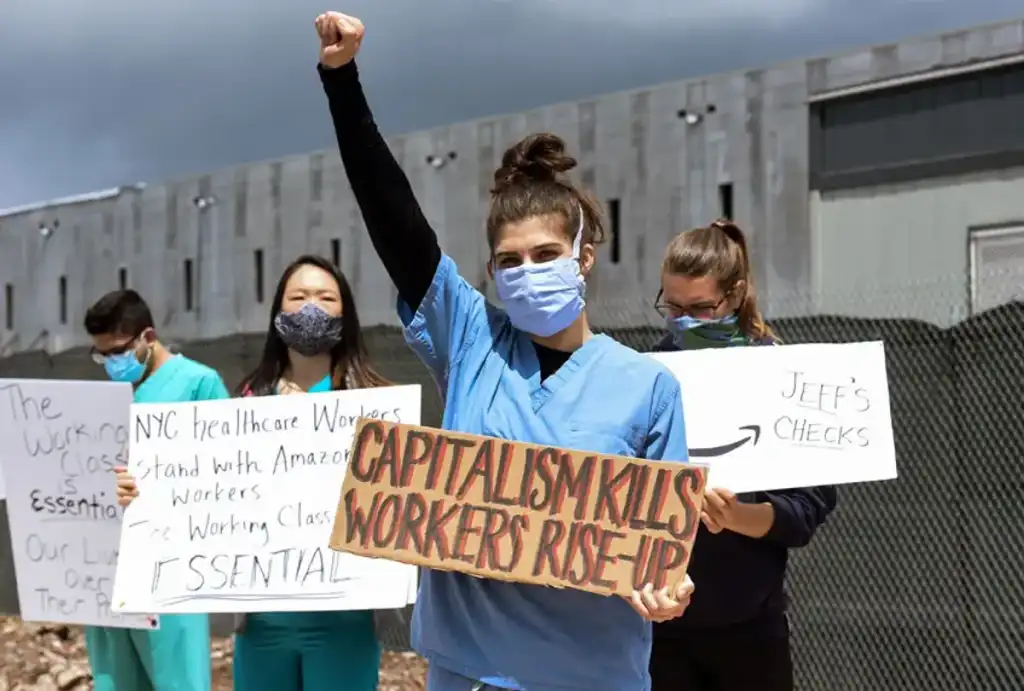 Four people in nurses scrubs holding protest signs. A women at the front has her fist raised and a sign that reads 'CAPITALISM KILLS WORKERS RISE-UP'